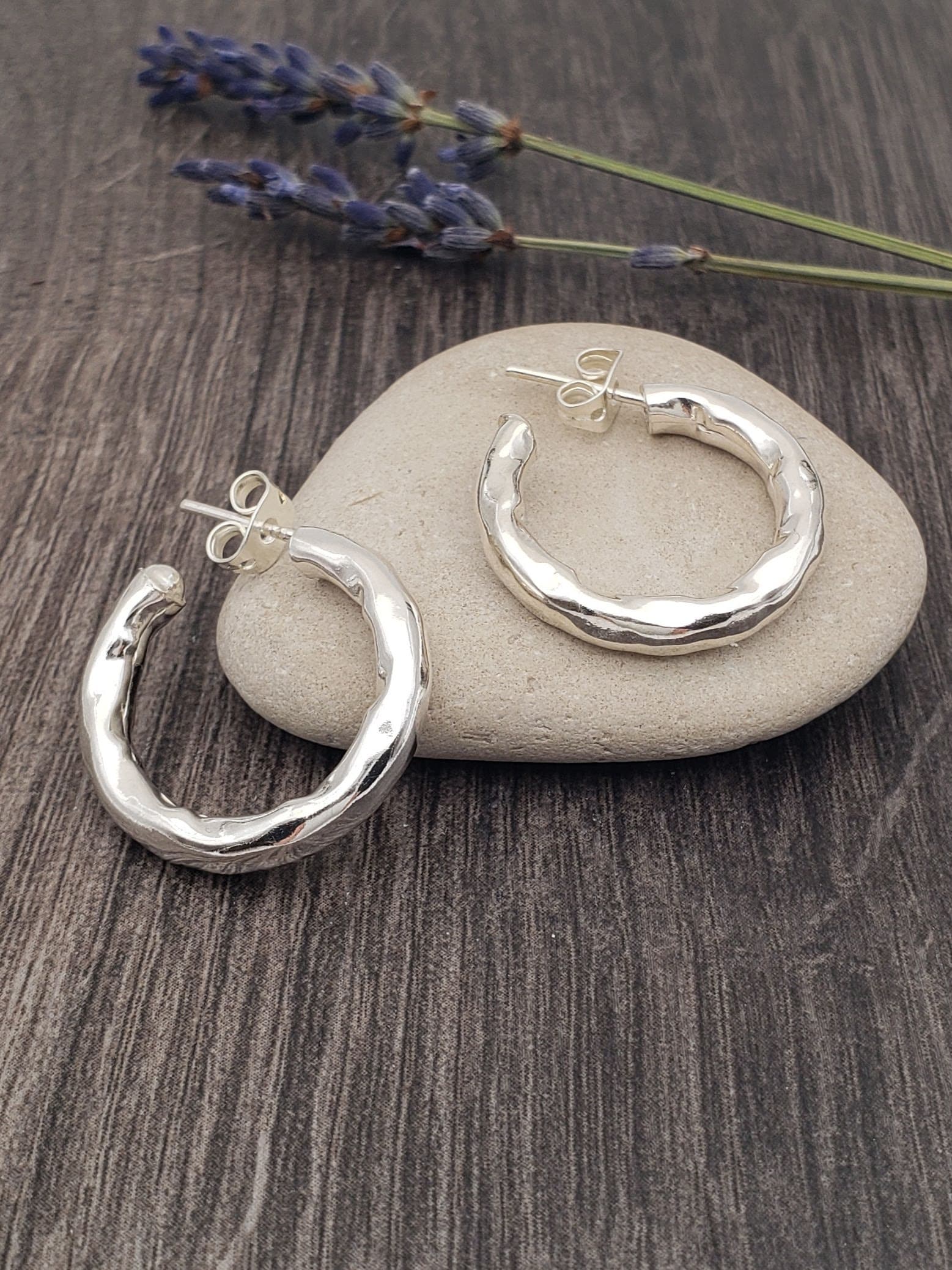 A pair of hammered hoop earrings placed on a smooth rock, with a lavendar sprig in the background